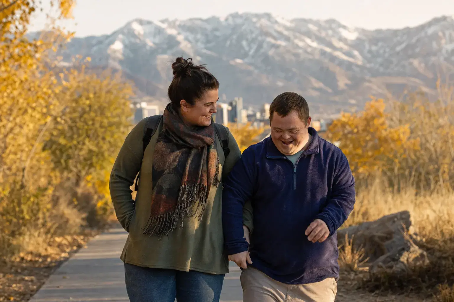 A direct support professional and an adult with a developmental disability walking a park trail with the Wasatch mountains behind them